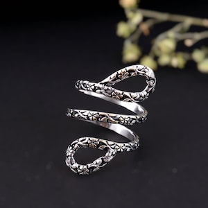 Könnte beinhalten: Ein silberfarbener Wickelring mit floralem Muster. Der Ring windet sich um den Finger und hat zwei schlaufenförmige Enden und eine strukturierte Oberfläche. Der Ring ist vor einem dunklen Hintergrund platziert.