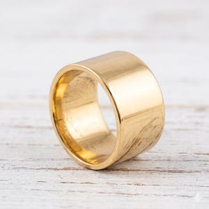 May include: A wide, gold-colored ring with a smooth, polished surface. The ring has a cylindrical shape and a simple, minimalist design. The ring is likely made of a precious metal, such as gold or gold-plated material.