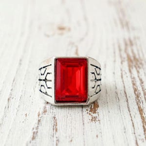May include: Silver-toned ring with a large, rectangular, bright red gemstone. The ring has a decorative pattern on the sides. The ring is set against a white wooden surface.
