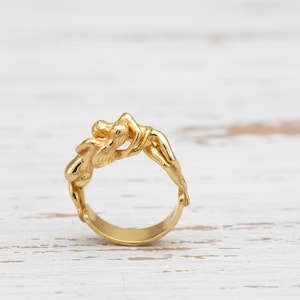 Op de afbeelding: Een goudkleurige ring met verweven menselijke figuren. Het ontwerp van de ring toont gebeeldhouwde vormen, waardoor een uniek en artistiek sieraad ontstaat. De ring is geplaatst op een lichtgekleurd houten oppervlak.