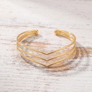 Puede incluir: Una pulsera tipo brazalete de tono dorado con tres bandas paralelas que convergen en un punto central en forma de V. El diseño abierto incluye pequeños terminales esféricos en cada extremo del alambre metálico, creando un aspecto geométrico.