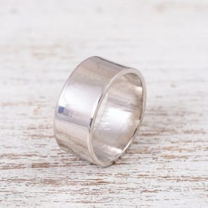 Op de afbeelding: Een brede, gepolijste zilverkleurige ring met een glad, reflecterend oppervlak. De ring heeft een eenvoudig, onversierd ontwerp met een constante breedte en vertoont een metaalachtige glans die het licht op de gebogen buitenkant opvangt.
