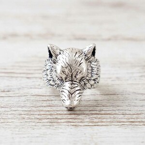 May include: A silver-colored ring featuring a detailed wolf head design. The ring showcases the wolf's facial features, including its ears, eyes, and snout. The ring is made of a shiny metal and is set against a light-colored wooden surface.