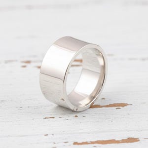 Könnte beinhalten: Ein glänzender, silberner Ring mit einem flachen, breiten Band. Der Ring hat eine glatte, polierte Oberfläche und ein schlichtes, modernes Design. Der Ring ist wahrscheinlich aus Metall und für den täglichen Gebrauch geeignet.