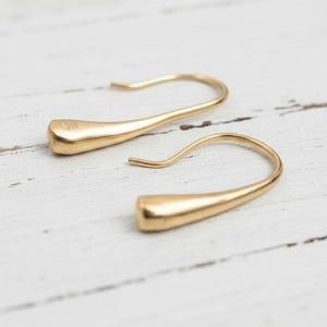 May include: A pair of gold-colored, teardrop-shaped earrings. The earrings have a curved hook design and a smooth, polished finish. The earrings are displayed on a white, textured surface.