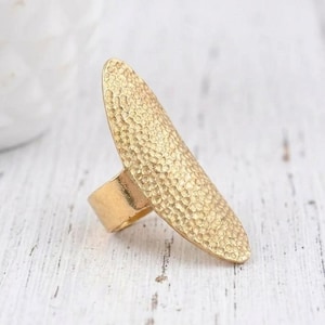 Può includere: Un anello color oro con un design ovale allungato e strutturato. L'anello ha un aspetto di metallo martellato e una fascia larga. Questo anello importante è adatto per aggiungere un tocco di eleganza a qualsiasi outfit.