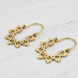 Può includere: Un paio di orecchini color oro con un design traforato e intricato. Ogni orecchino presenta una forma a semicerchio con una serie di ritagli decorativi e una semplice chiusura a gancio. Gli orecchini sono esposti su una superficie di legno bianco.