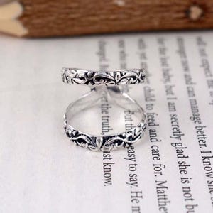 Può includere: Un anello argentato dal design aperto, con intricati motivi floreali e a volute. L'anello ha due fasce collegate da un anello centrale, creando un look unico ed elegante. L'anello è posto su uno sfondo di testo stampato.