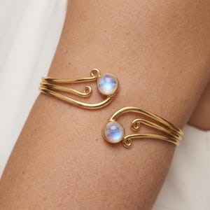May include: A gold-toned open cuff bracelet featuring three parallel wire bands that curve into decorative swirls at each end. Each terminal is accented with a round, iridescent moonstone cabochon, creating a symmetrical design.