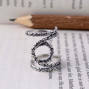 May include: A silver-colored ring with a spiral design. The ring features a floral pattern and is set against a backdrop of a book page and a pencil.
