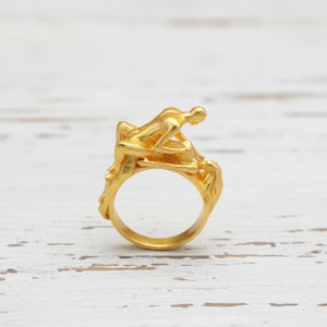 Puede incluir: Un anillo dorado que presenta dos figuras humanas estilizadas en un abrazo sensual. El diseño del anillo incluye detalles intrincados, con las figuras situadas sobre una banda texturizada. El anillo está sobre una superficie de madera blanca desgastada.