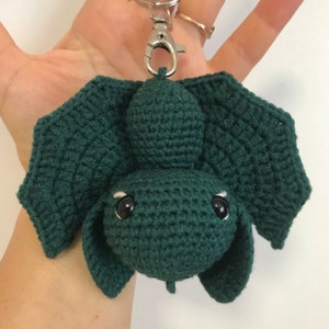 May include: A green crocheted bat keychain with black eyes. The bat has a small, round head and large, pointed wings.