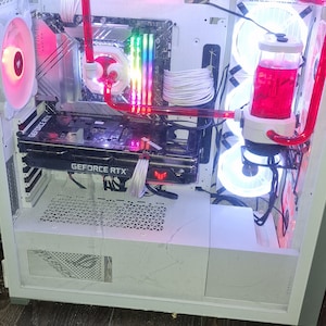 Pode incluir: Uma caixa de computador branca com componentes internos, incluindo uma placa gráfica com a etiqueta "GEFORCE RTX" e um sistema de refrigeração líquida com tubos vermelhos e um reservatório. A caixa tem múltiplos ventiladores com iluminação LED.