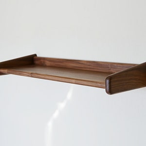 May include: A dark brown wooden wall shelf with rounded edges. The shelf is mounted to a white wall.