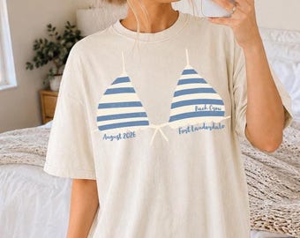 May include: A cream-colored short-sleeve t-shirt featuring a graphic of two blue and white striped bikini tops. The text "August 2026" is printed below the left cup, and "Bach Crew Fort Lauderdale" is printed below the right cup in blue script.