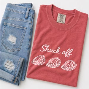 May include: A light blue denim jeans with distressed details and a red t-shirt. The t-shirt has the text "Shuck off" with three white oyster illustrations. The t-shirt is a Comfort Colors brand.
