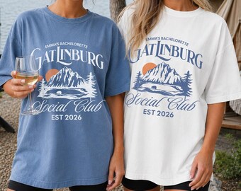 May include: Two crewneck t-shirts, one blue and one white, featuring a graphic print of a mountain landscape, a sun, and trees. The text reads: Emma's Bachelorette Gatlinburg Social Club EST 2026. The shirts have short sleeves and a relaxed fit.