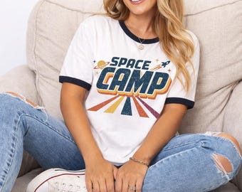 May include: A white ringer t-shirt with dark blue trim on the collar and sleeves. The front features the text SPACE CAMP in bold blue letters with a yellow and orange outline, accompanied by a small rocket, a planet, and colorful sun rays.