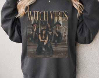 May include: A charcoal gray crewneck sweatshirt featuring a rectangular graphic print of four individuals in witch hats sitting on wooden porch steps. The text WITCH VIBES is printed in a tan serif font above the photographic image of the group.