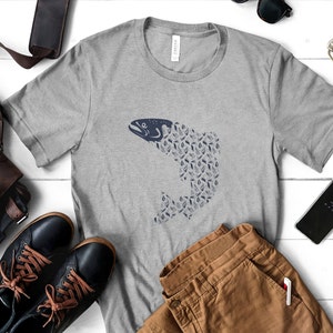 May include: A grey t-shirt featuring a blue fish design. The fish is made up of smaller blue shapes resembling leaves. The shirt is laid flat on a white surface with brown trousers, a pair of black shoes, and a mobile phone.