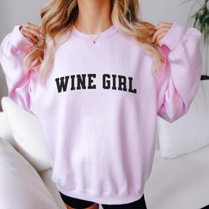 Pu&ograve; includere: Una felpa rosa con la scritta "WINE GIRL" stampata in nero.