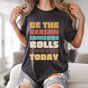 May include: A dark gray t-shirt with the text "BE THE REASON SOMEONE ROLLS THEIR EYES TODAY" in colorful vintage-style lettering.