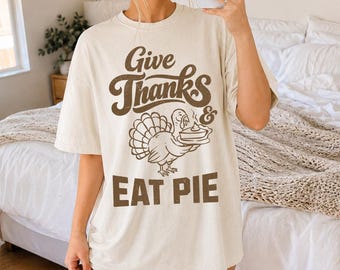 May include: A cream-colored short-sleeve t-shirt featuring brown screen-printed text that reads Give Thanks & EAT PIE. The graphic includes a central illustration of a turkey holding a pie on a platter, rendered in a simple line-art style.
