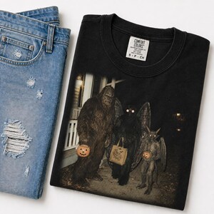 Puede incluir: Una camiseta negra de cuello redondo con un estampado gráfico de Bigfoot, una criatura alada y un pequeño demonio con cuernos de pie juntos por la noche, cada uno sosteniendo un cubo de calabaza, junto a unos vaqueros azules desgastados.
