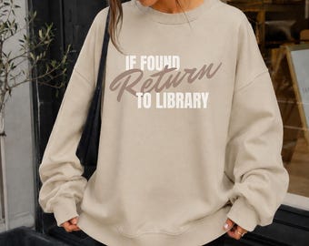May include: A beige crewneck sweatshirt featuring the white and brown text IF FOUND RETURN TO LIBRARY printed on the front. The garment has long sleeves with ribbed cuffs and a ribbed hem, providing a relaxed fit. A black shoulder bag strap is visible.