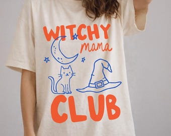May include: A cream-colored t-shirt featuring the orange text "WITCHY mama CLUB" and blue line art graphics of a crescent moon, small stars, a cat, and a pointed hat. The design is centered on the front of the short-sleeved garment.
