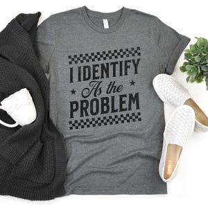 Puede incluir: Una camiseta gris jaspeado con el texto "I Identify As the Problem" en negro. La camiseta se combina con un cárdigan gris oscuro, una taza blanca y zapatos sin cordones blancos. También se ve una pequeña planta verde.