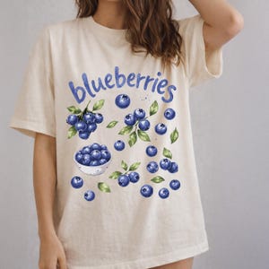 Op de afbeelding: Crèmekleurig T-shirt met een aquarelontwerp van blauwe bessen en groene bladeren. Het woord "blueberries" staat in blauwe letters bovenaan. Een kom met blauwe bessen is ook in het ontwerp verwerkt.