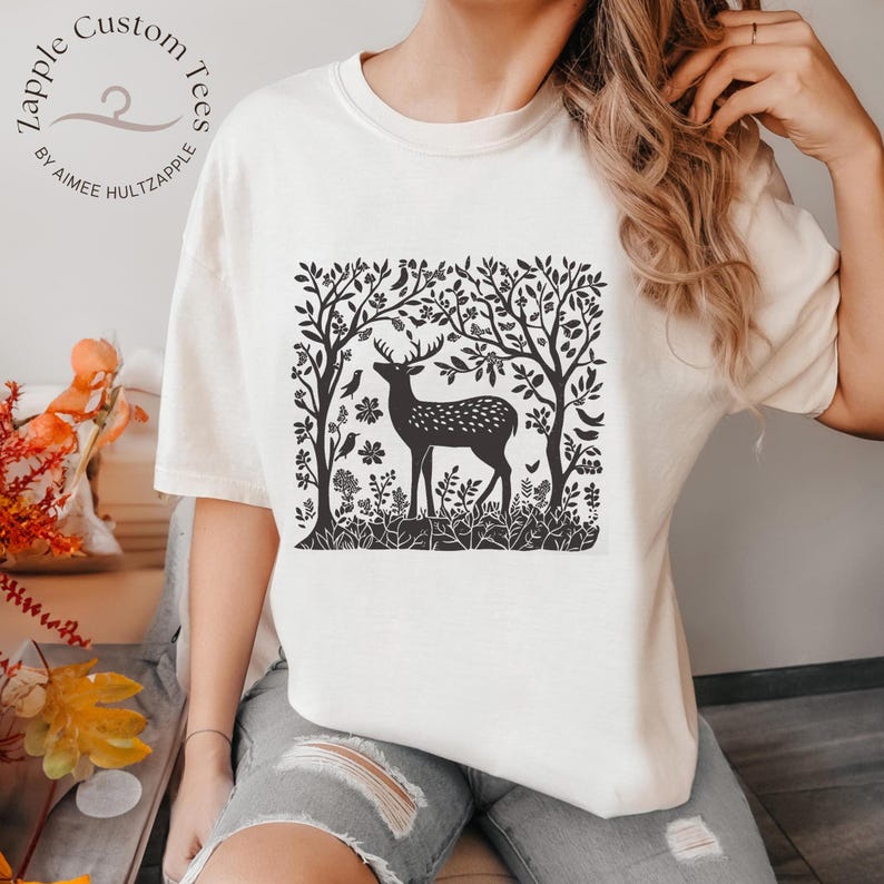 K&ouml;nnte beinhalten: Ein wei&szlig;es T-Shirt mit einem schwarz-wei&szlig;en Grafikdruck eines Hirsches in einem Wald. Die Grafik zeigt einen Hirsch, der in einem Wald mit B&auml;umen, V&ouml;geln und Blumen steht. Der Text "Zapple Custom Tees by Aimee Hultzapple" ist oben links auf dem Shirt gedruckt.