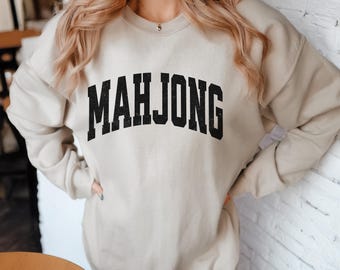 May include: A beige crewneck sweatshirt featuring the word MAHJONG printed in a large, arched, black, distressed-style font across the chest. The garment has long sleeves, a ribbed neckline, and a relaxed fit, providing a simple, neutral look.