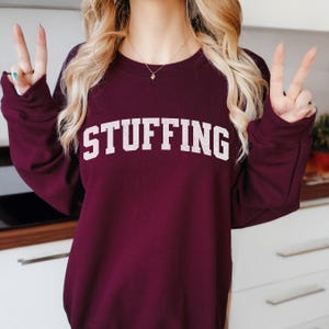 May include: A burgundy sweatshirt with the word "STUFFING" printed in large, white, block letters across the chest. The sweatshirt is a solid color and has long sleeves. The image shows a person wearing the sweatshirt.