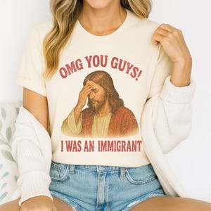 May include: Cream-colored t-shirt with the text "OMG YOU GUYS! I WAS AN IMMIGRANT" in red. The shirt features a distressed image of Jesus with his hand on his forehead. The shirt is paired with blue denim shorts.