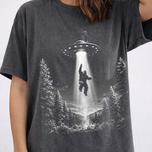 Può includere: Una t-shirt grigio antracite con una stampa grafica che raffigura una creatura Bigfoot rapita da un disco volante. La scena mostra un paesaggio boschivo con pini e un raggio di luce brillante che scende dall'UFO verso la creatura.