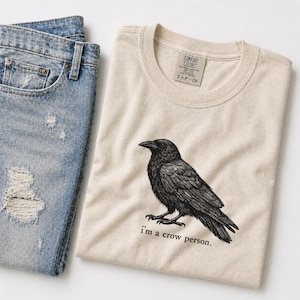 May include: A cream-coloured t-shirt featuring a black crow illustration and the text "I'm a crow person." Light blue denim jeans with distressed details are next to the shirt. The shirt has a Comfort Colors tag.