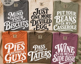May include: Six crew neck t-shirts in charcoal, cream, olive green, orange, brown, and plum. Each shirt features white or black screen-printed text with phrases including Mind Your Own Biscuits, Just One More Deviled Egg, and Wine Is My Side Dish.