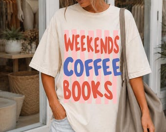 May include: An off-white short-sleeve t-shirt featuring vertical light pink stripes and the text "WEEKENDS COFFEE BOOKS" in red and blue lettering. The shirt has a crew neckline and a loose fit, paired with light blue denim shorts and a large gray tote bag.