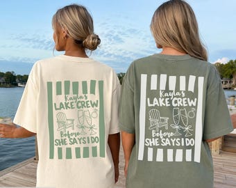 May include: Two short-sleeve t-shirts, one ivory and one moss green, each featuring a back graphic with vertical stripes, a chair, two champagne flutes, and a ring icon. The text reads: Kayla's LAKE CREW Before SHE SAYS I DO.