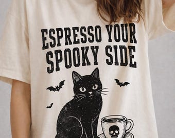 May include: A cream-colored t-shirt featuring a black graphic print of a cat sitting next to a coffee cup with a skull design. Above the cat, the text reads "ESPRESSO YOUR SPOOKY SIDE" in a bold, distressed black font, accompanied by three small bats.