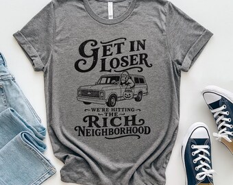 May include: A heather gray short-sleeve t-shirt featuring a black graphic of a gorilla driving a pickup truck with a pumpkin. The text reads: GET IN LOSER WE'RE HITTING THE RICH NEIGHBORHOOD. The shirt is displayed alongside blue jeans and blue sneakers.