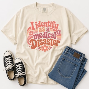 Może przedstawiać: Kremowy t-shirt z krótkim rękawem z napisem "I identify as a medical DISASTER" wykonanym retro, zakrzywioną czcionką. Wzór zawiera różowe, pomarańczowe i czerwone łuki oraz małe akcenty w kształcie kwiatów i gwiazdek nadrukowane z przodu.