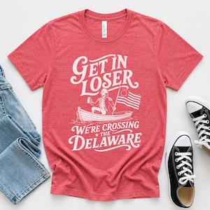 May include: A heather red short-sleeve t-shirt featuring a white graphic of a person in a boat with an American flag and the text "GET IN LOSER WE'RE CROSSING THE DELAWARE" printed in a stylized font across the center of the chest.