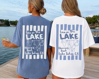 May include: Two oversized t-shirts, one blue and one white, featuring a back graphic with vertical stripes and the text Love on the LAKE Hanna's Bach Lake Tahoe, CA, accompanied by illustrations of a bikini and a diamond engagement ring.