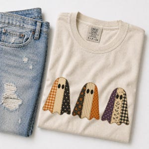 May include: A beige crew neck t-shirt featuring a graphic of three ghosts made from various patterned fabric swatches, including plaid, floral, and star prints. The shirt is paired with a pair of distressed blue denim jeans with visible wear.