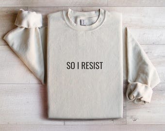 Può includere: Una felpa beige chiaro con la scritta "SO I RESIST" stampata in nero sul davanti.