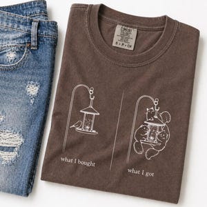 Könnte beinhalten: Braunes T-Shirt mit weißer Grafik eines Vogelfutterhäuschens. Der Text "what I bought" steht unter dem Vogelfutterhäuschen links, und "what I got" steht unter einem mit Eichhörnchen gefüllten Futterhäuschen rechts. Zerrissene blaue Jeans im Hintergrund.