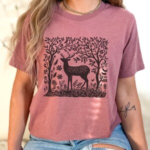 K&ouml;nnte beinhalten: Ein mauvefarbenes T-Shirt mit einem schwarz-wei&szlig;en Grafikdruck eines Hirsches in einer Waldszene. Die Grafik zeigt einen Hirsch mit Geweih, B&auml;ume und V&ouml;gel.